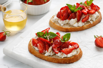 Sweet Toast with cottage cheese ricotta cheese, strawberry, honey and mint. Sandwiches on white marble board and paper. Bruschetta, snack, appetizers.