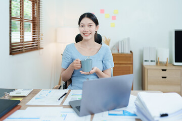 Portrait of an Asian business woman drinking coffee while working with a computer and financial statements documents on her desk