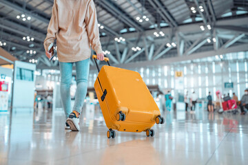 Young female traveler passenger walking with a yellow suitcase at the modern Airport Terminal, Back...