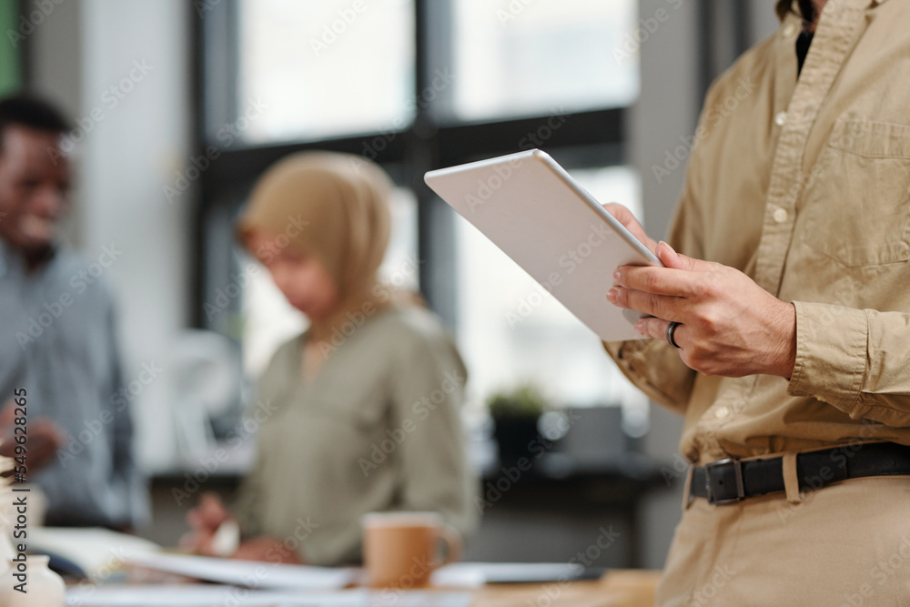 Aufkleber Hands of young analyst or broker using digital tablet while standing in front of camera against colleagues discussing points of working plan