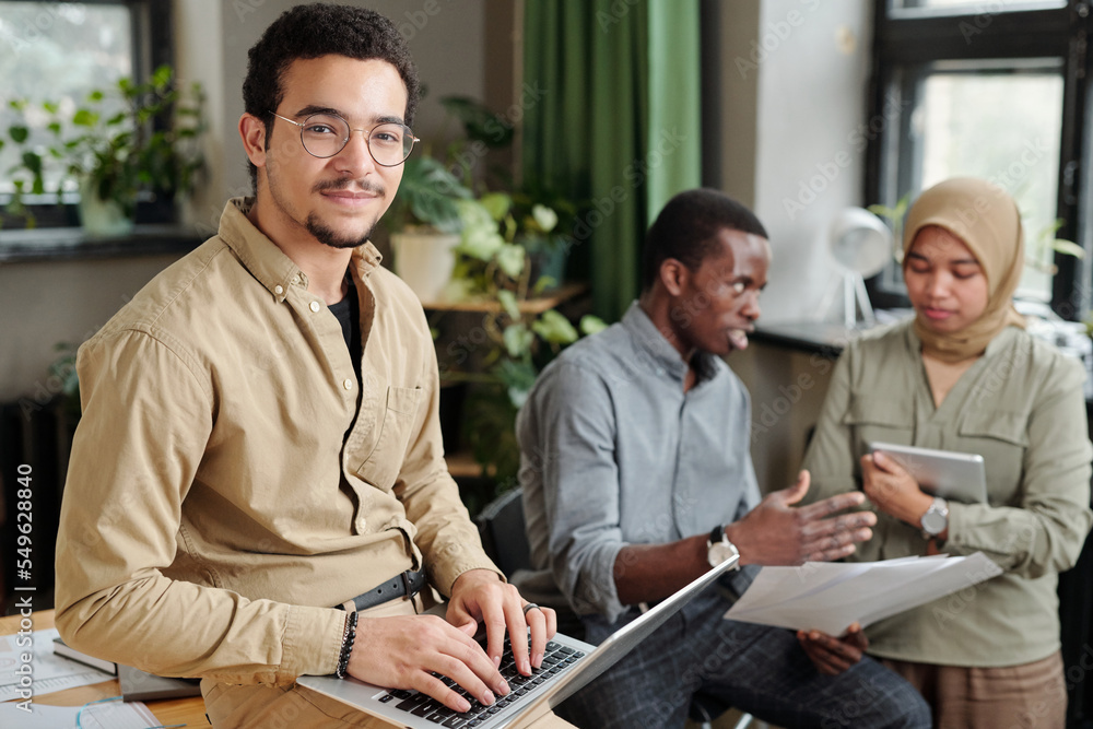 Poster young confident broker looking at camera while sitting on workplace against two intercultural cowork