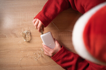Female Santa Claus charge garland with power bank at table, top view. Power crisis problem
