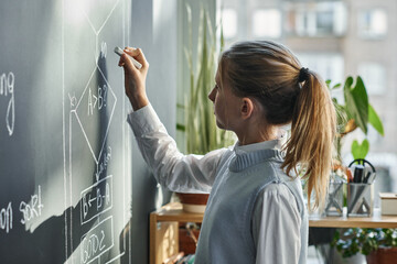 Schoolgirl writing formula on blackboard with chalk during lesson at classroom