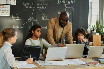 African American teacher teaching students to use computers, they discussing software sitting at...