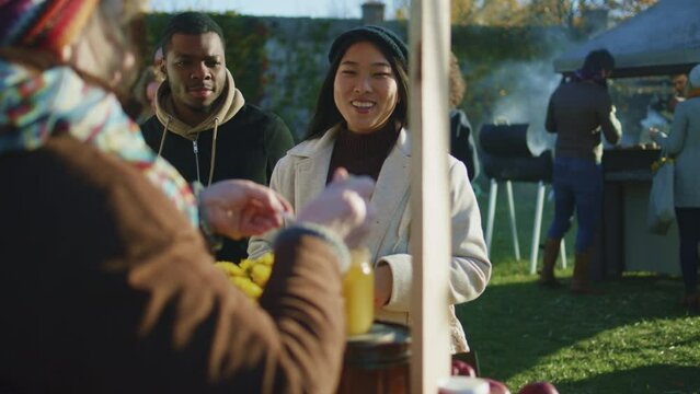 Diverse couple shopping at local farmers market. Asian woman tastes a spoon of honey. People walking on background. Autumn fair on weekend outdoors. Vegetarian and organic food.