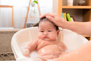 Asian newborn baby in bathtub taking a shower filled with soap bubbles, single mother bathing daughter and washing baby head with warm water with carefully and love, hygiene concept.