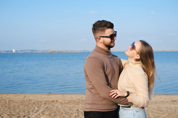 Fototapeta premium A young couple in jeans, sweaters and sunglasses stands on the beach near the lake and hugs