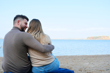 Fototapeta premium A young man and woman are sitting against the backdrop of a large blue lake and hugging. couple sitting with their backs to the camera