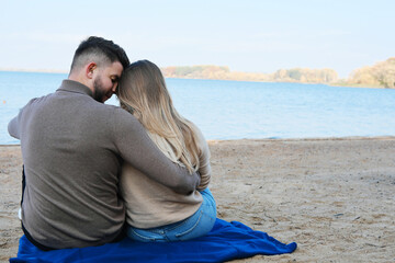 A young man and woman are sitting against the backdrop of a large blue lake and hugging. couple sitting with their backs to the camera