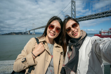 two cheerful asian Korean female friends wearing sunglasses and looking at camera with smiling face. they are having fun taking selfie pictures with Oakland bay bridge in san Francisco