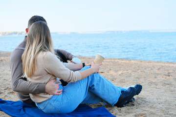 Date on the beach. A young couple in sweaters sits on a plaid, holds coffee in stacks in their hands and kisses.