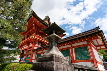 Kyoto, Japan - April 18, 2018: low angle shot of a toro stone lamp standing near the red temple building at Fushimi Inari Taisha shrine against blue sky - obrazy, fototapety, plakaty
