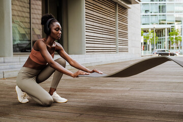 Black young sportswoman listening music and unrolling yoga mat