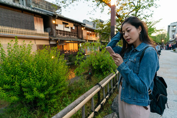 concentrated asian Japanese lady traveler pushing hair back while reading online travel info on phone at geisha district in gion Kyoto japan in the evening