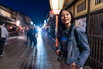 happy asian woman photographer looking around at nostalgic hanamikoji street lined with shops and geisha houses with red illuminated lamp post at background in gion Kyoto japan at night