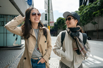 two smiling asian chinese female coworkers chatting happily on pedestrian walk while walking to...