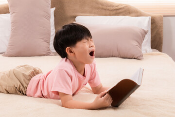 Asian little child boy feeling sleepy yawn and rubbing both eyes with both hands while reading favorite manga, elementary children tired after school lying on bed and read book, doing homework.