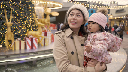 smiling asian mother talking to her baby in arms while they are looking at festive city scene near a water fountain with decorations celebrating Christmas holiday
