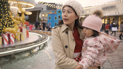 cheerful asian mother and her innocent baby daughter enjoying Christmas atmosphere near a decorated fountain on a city square in the evening