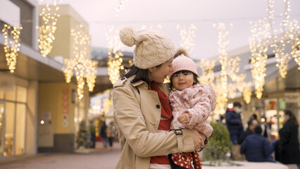 Fototapeta na wymiar cheerful asian mother talking to her baby while they are watching and pointing up at charming holiday light decorations outside shopping mall in Christmas season
