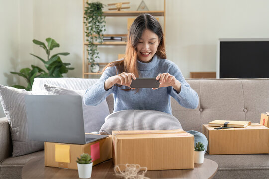 Young Asian Woman Take Photos To Box Addresses Smartphone For Post To Sell Online On The Internet And Prepare Pack Product Boxes. Selling SME Online Ideas Concept