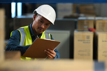 Asian male manager wearing hardhat and reflective jacket checking inventory in a warehouse with shelves full of cardboard boxes