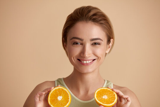 Laughing Girl Hold Slices of Orange. Happy Young Woman Looking at Camera and Recommended Vitamin for Skin. Concept of Skincare. Isolated on Beige Background 