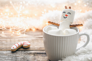 Close up, mug of hot drink with marshmallow snowman on blurred background.
