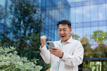 Asian businessman in casual clothes with phone outside office building celebrating victory and...