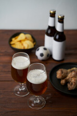 Two glasses with beer, bottles, snacks and soccer ball on wooden table.