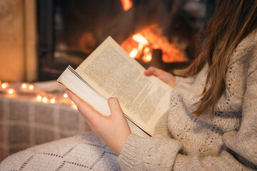 Obraz premium Little girl reading a book sitting by the fireplace at home, close-up.