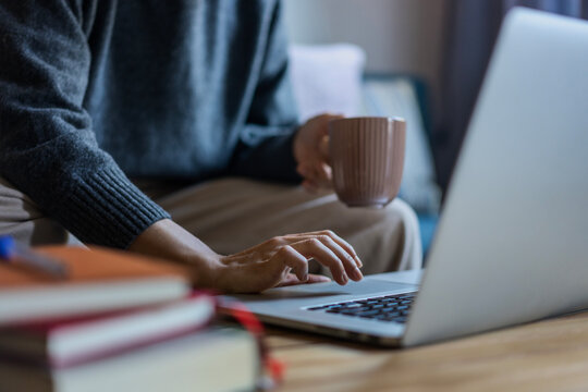 Closeup shot of a female hand working on a laptop using a touchpad. A woman in a warm sweater holds a mug of coffee in her other hand