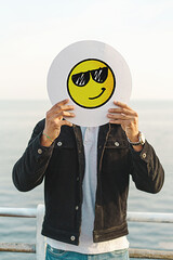 Young adult man cover face with a smiling emoji face with sunglasses, hand drawn on a white disc