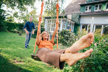 Happy woman swinging on swing with man standing behind in back yard
