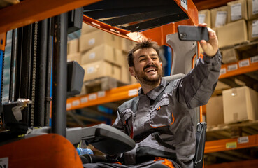 Young wearhouse forklift worker, having small break from work and using phone while sitting on forklift, taking selfies