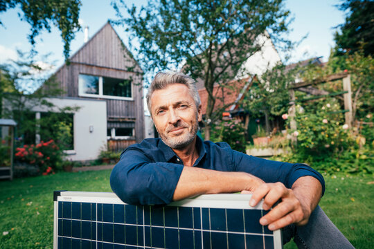 Smiling mature man sitting with solar panel in front of house