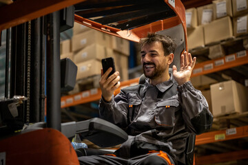 Young wearhouse forklift worker, having small break from work and using phone while sitting on forklift, taking selfies