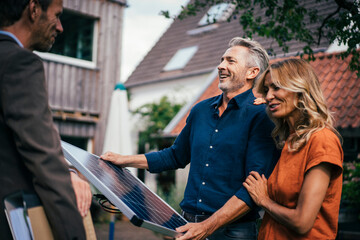 Happy couple holding solar panel and discussing with real estate agent in back yard