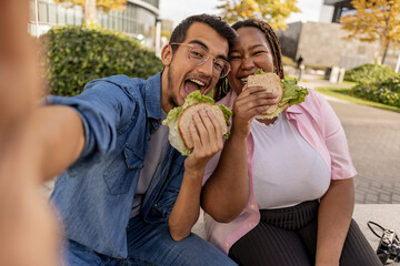 Happy young couple eating sandwich and taking selfie at footpath