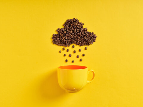 Raining coffee concept. Coffee beans in shape of rain cloud and raindrops raining towards the coffee cup on yellow background.