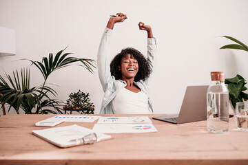 Happy businesswoman with arms raised at desk in home office