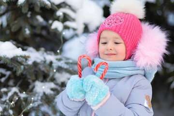 Cute little girl with candy cane next Christmas tree outdoor. Happy child waiting present for new year. Kid hands in snow blue mittens holding gift for xmas