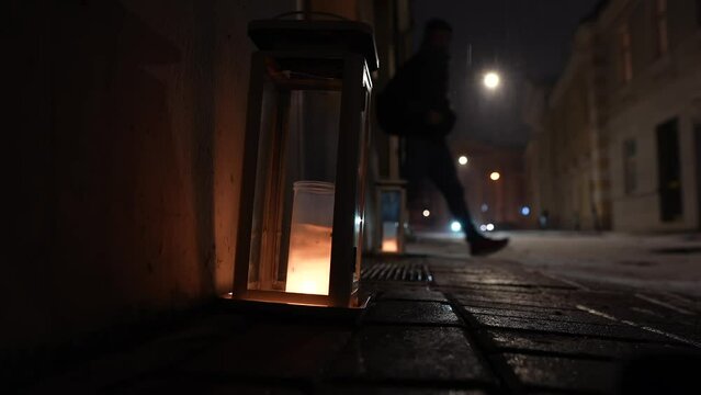 Candle light lamp placed at the corner on the street during a winter evening with calm snow falling on top of it and people silhouette in background. 4k concept video for winter holidays.