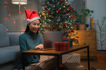 Happy Asian woman with santa hat using laptop for shopping or chat with friends during Christman holiday at home