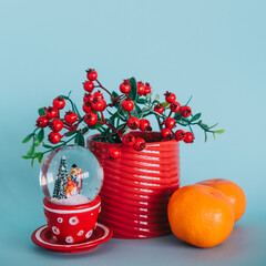 Snow globe with a branch of winter berries in a red mug and two tangerines on a blue background