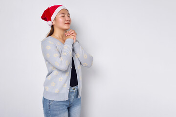 Hopeful young Asian woman in Christmas hat keeps palms pressed together in praying gesture isolated...
