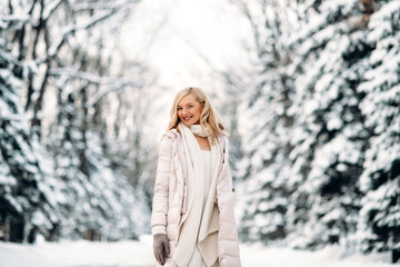 Fashion young smiling blonde woman in winter. Standing among snowy trees in winter forest. Wearing beige coat, white scarf in boho style