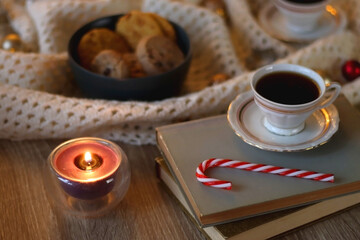Cups of tea or coffee, bowl of cookies, soft blanket, lit candles, books and various Christmas decorations on the table. Selective focus.