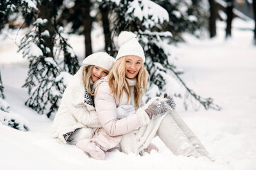 Fototapeta na wymiar Handsome mother and daughter are having fun outdoor in winter time. Sitting on snow in forest between snowy trees