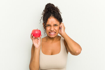 Young brazilian woman holding a red apple isolated covering ears with hands.